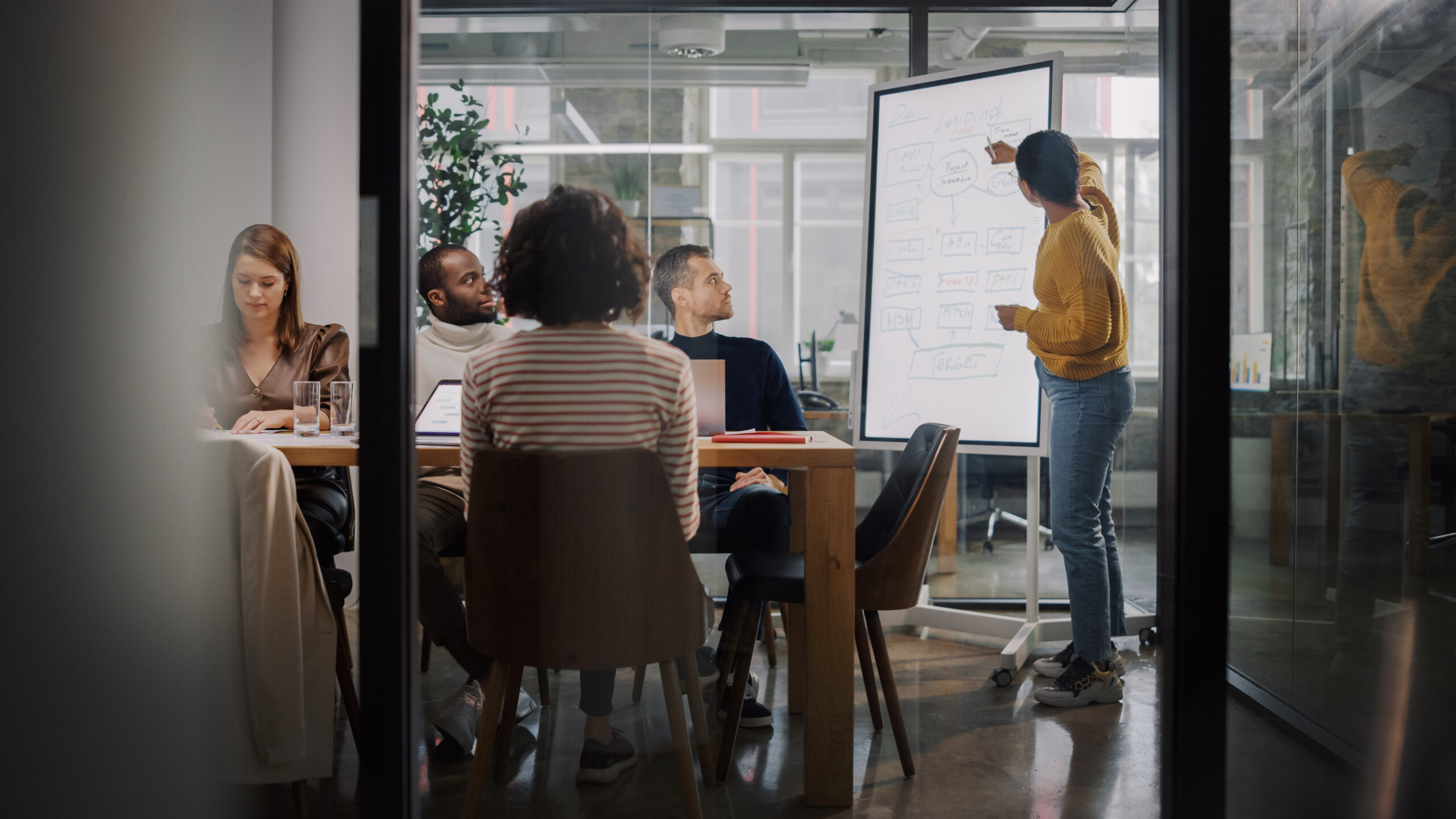 Project Manager Makes a Presentation for a Young Diverse Creative Team in Meeting Room in an Agency. Colleagues Sit Behind Conference Table and Discuss Business Development, User Interface and Design.